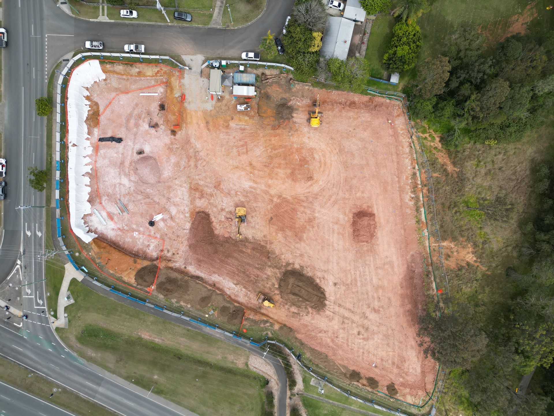 Aerial view of a cleared construction site with orange soil. A few machines and temporary structures are present.