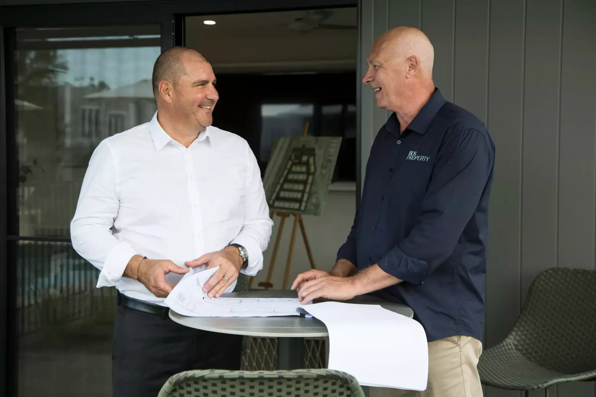 Two men, one in a white shirt, the other in navy, smile while looking at plans outdoors.