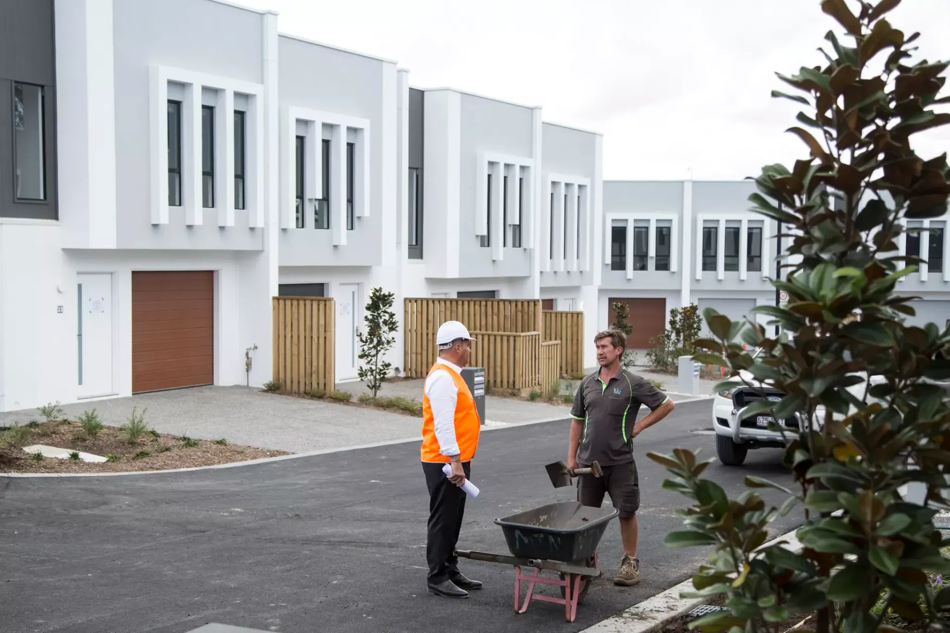 Two men discussing plans on a new housing development. One holds a wheelbarrow; gray buildings in background.