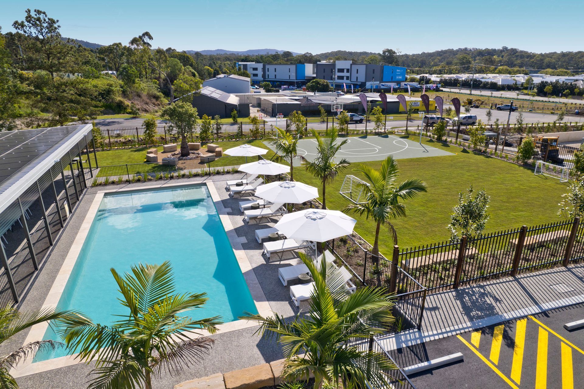 Pool area with umbrellas, green grass, and palm trees, set in front of buildings.