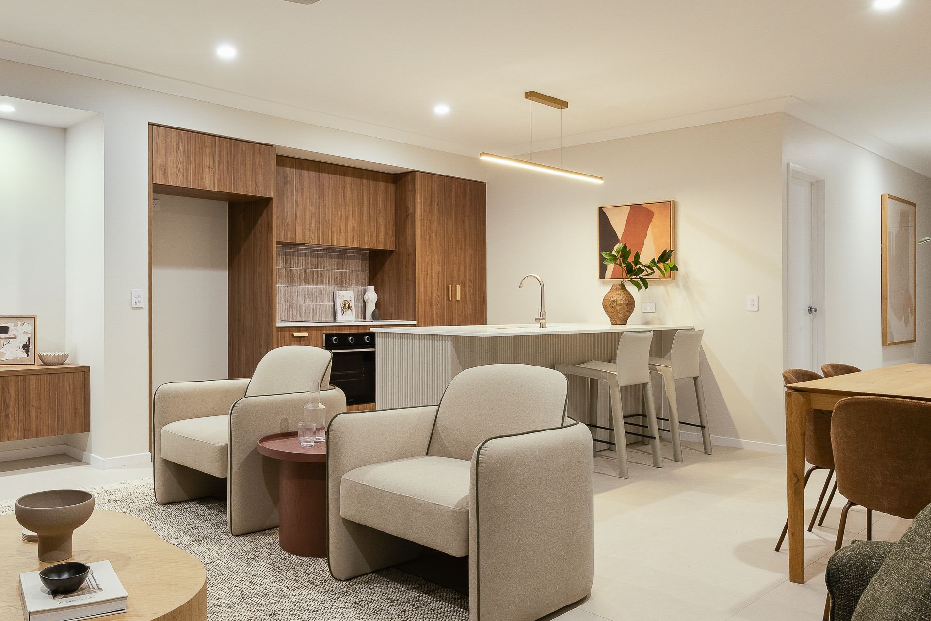 Modern open-plan living room and kitchen with beige armchairs, dining table, wood cabinetry, and warm lighting
