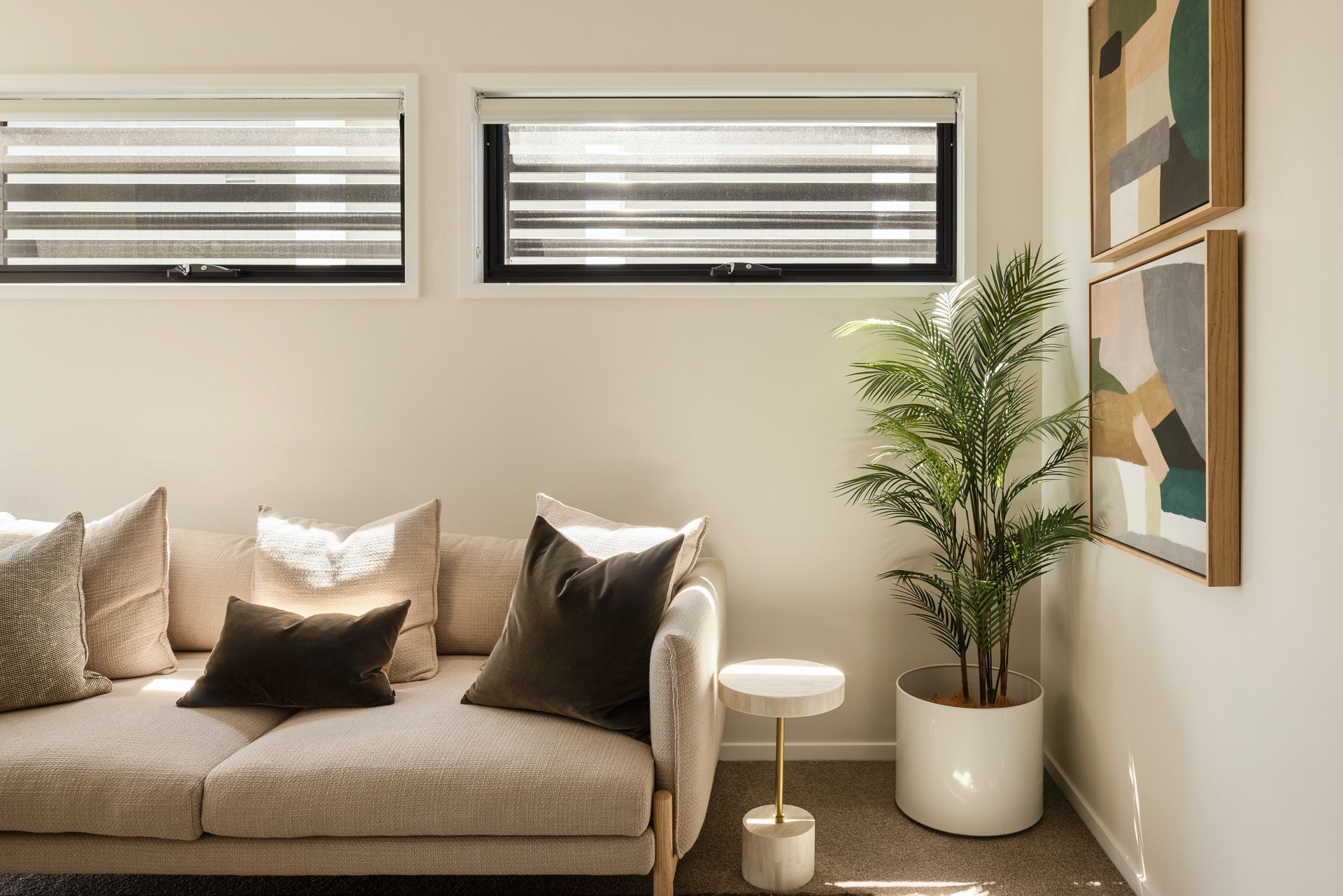Cozy beige sofa with pillows in a sunlit living room, beside a potted plant and wall art