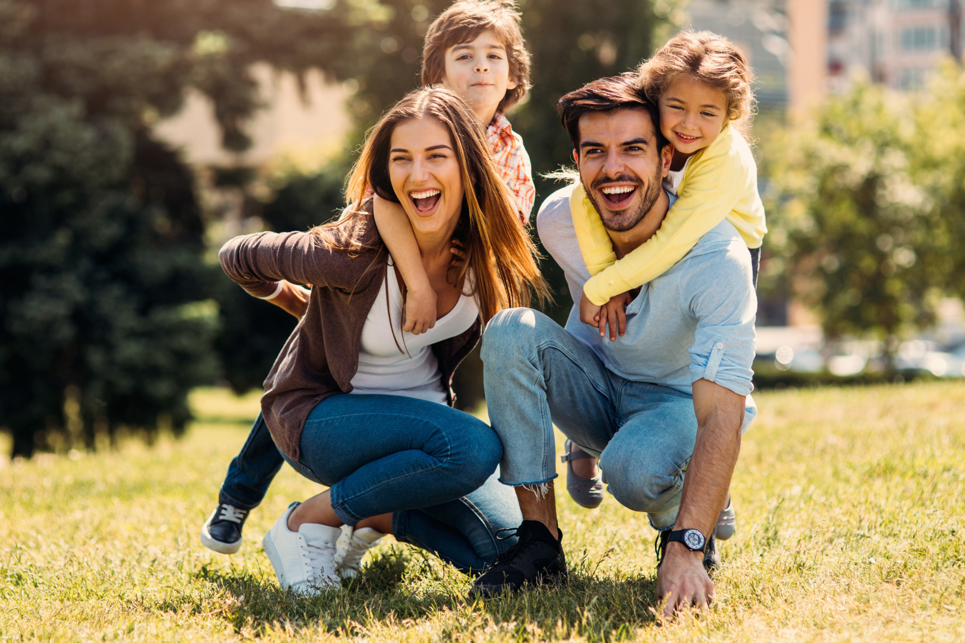 Family of four playfully posing in a grassy park, smiling and enjoying the sunlight.