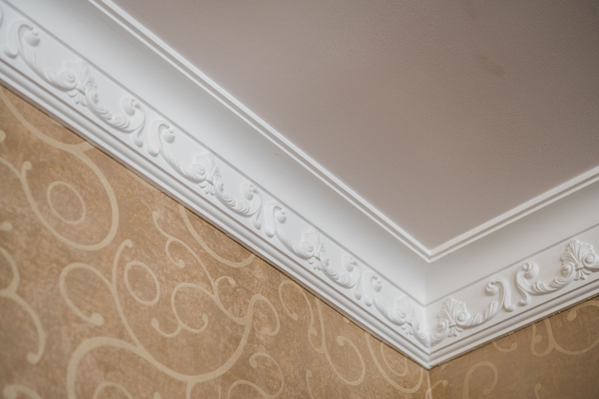 Corner of a room showing ornate white crown molding against a beige patterned wallpaper and a white ceiling.