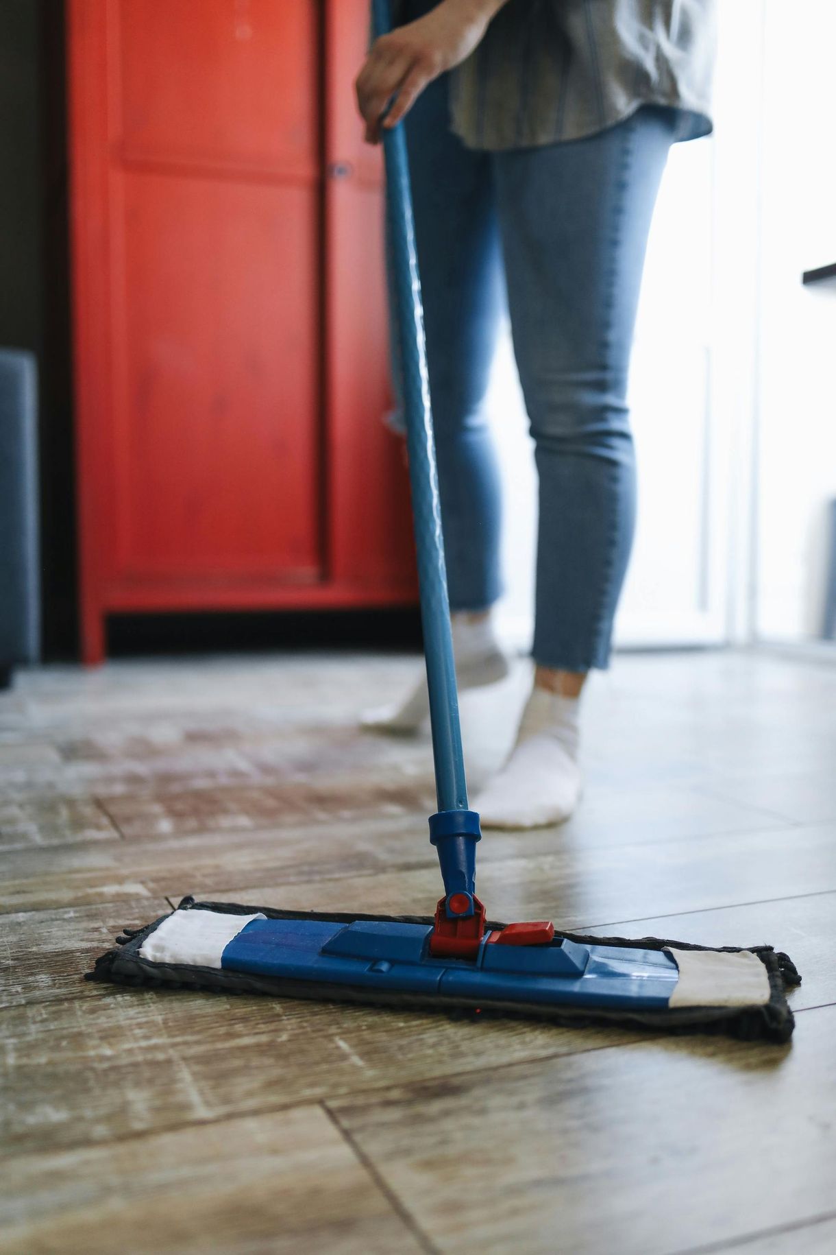 Person mopping a light wood floor with a blue mop; red cabinet in the background.
