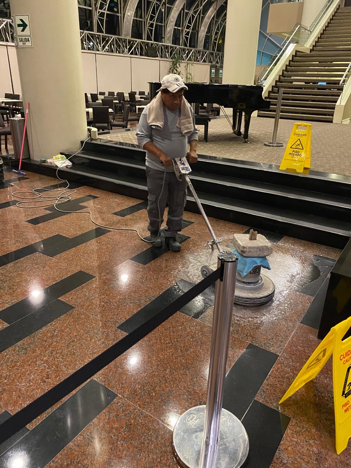 A worker polishes a floor in a building, using a machine and wearing a hat. Caution signs are visible.