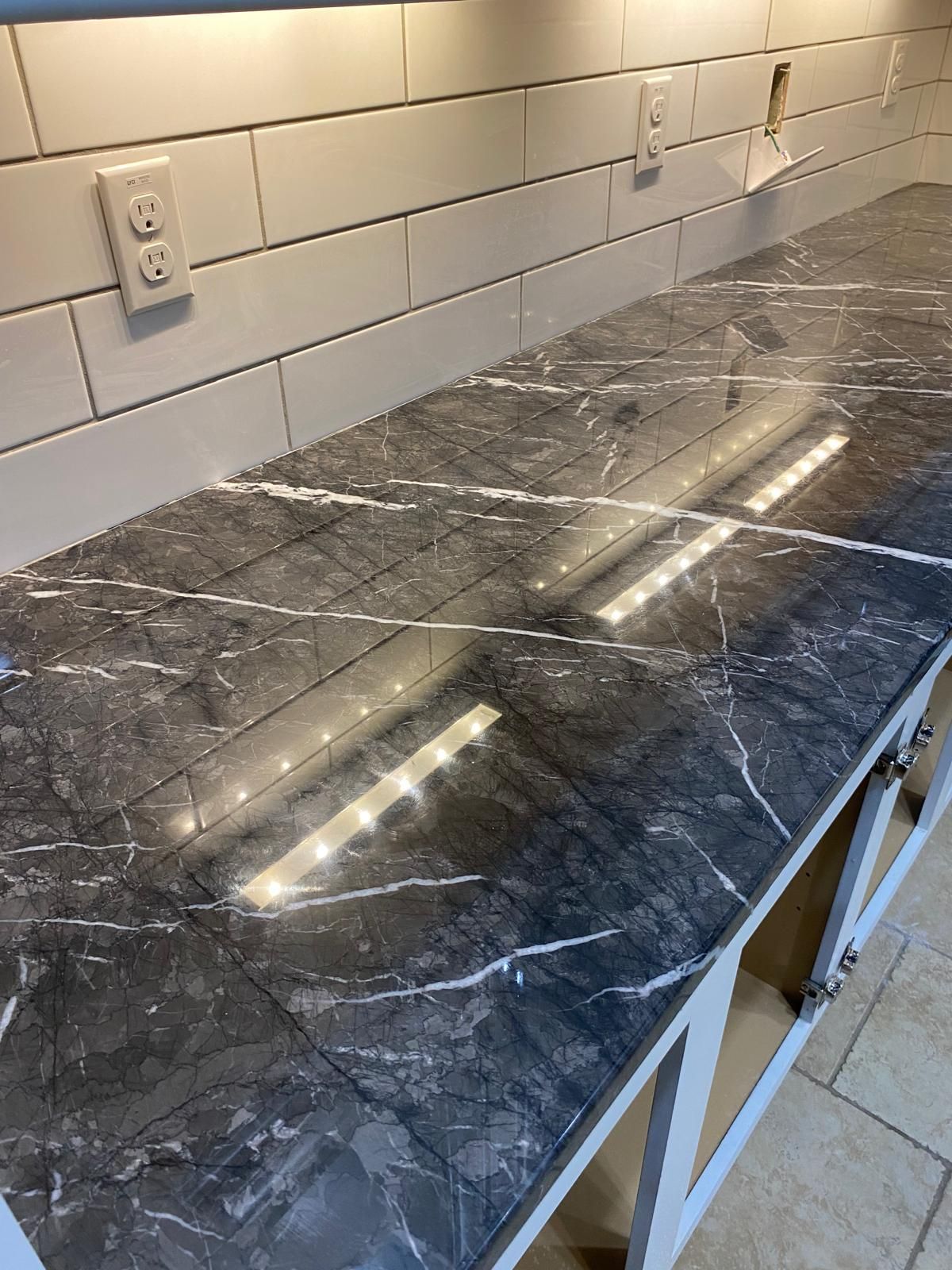 Dark countertop with white veining, under-cabinet lighting, white subway tile backsplash, and unfinished cabinets.