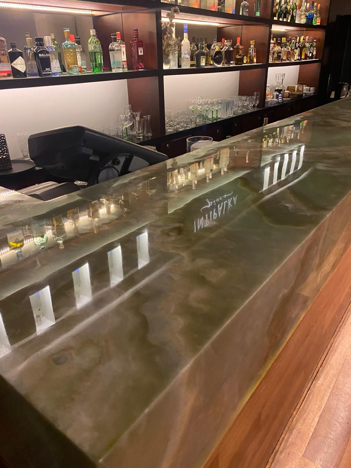 A glossy green bar counter reflects the bar's interior. Shelves stocked with liquor bottles are visible.