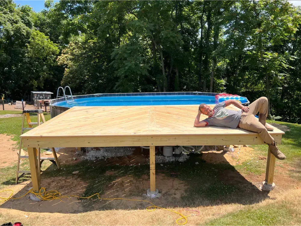 Man resting on new wooden deck near a pool. Green trees in background, sunny day.