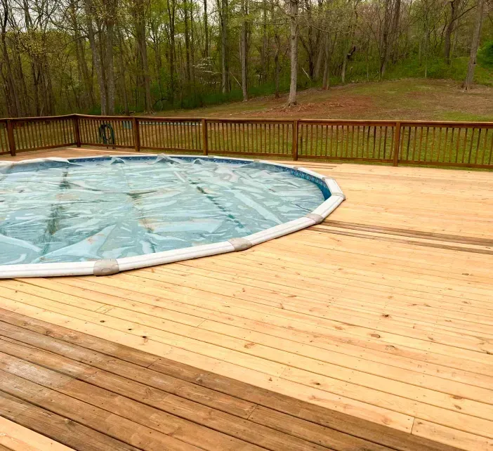 A deck surrounds a round pool covered in blue plastic. A wooden fence borders the deck with trees in the background.