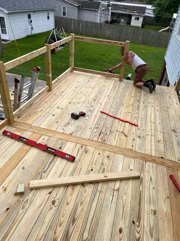 Person building a wooden deck outdoors; deck boards laid, railing partially complete.
