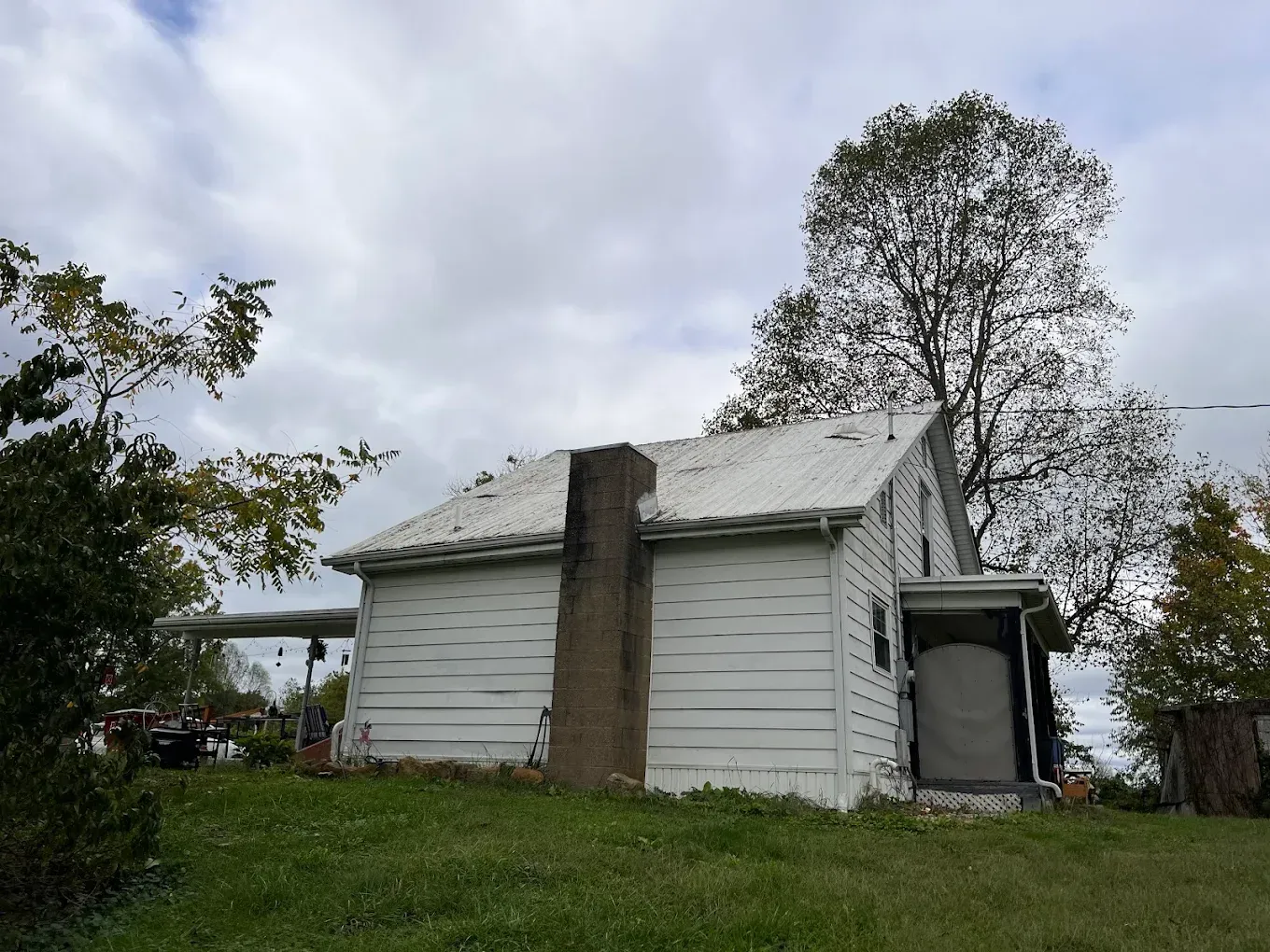 White house with chimney, porch, and overgrown yard on a cloudy day.