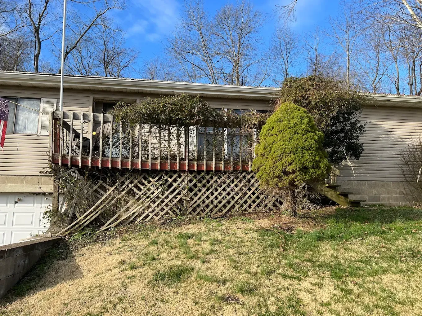 A house with a weathered wooden deck, surrounded by sparse grass and bare trees under a blue sky.