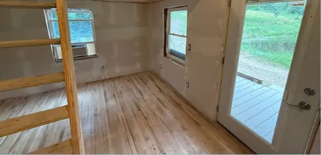 Interior of a tiny house with wood floors and ladder to a loft. Door opens to outdoor greenery.