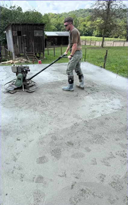 Man using a concrete finishing machine on a concrete slab outdoors, near a wooden shed and trees.