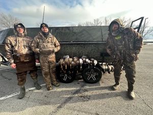 Three men are standing in front of a boat filled with ducks.