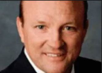 Headshot of a smiling person wearing a dark suit jacket and white shirt against a neutral grey background.