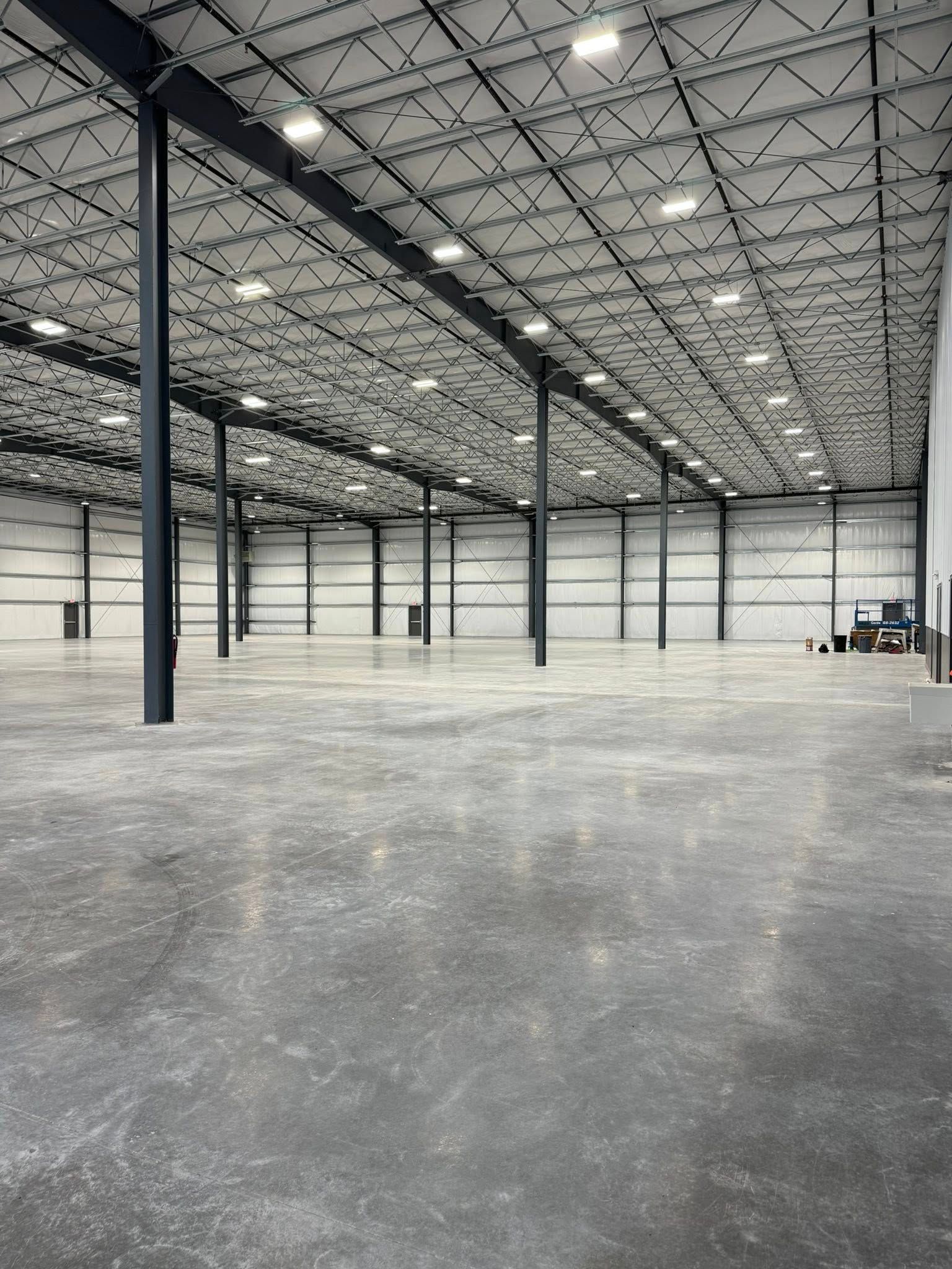 A spacious, empty industrial warehouse with a high, exposed truss ceiling, polished concrete floor, and black steel beams.