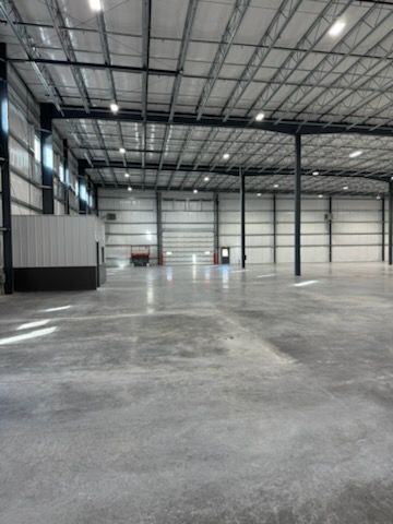 A spacious, empty industrial warehouse with a high steel-truss ceiling, concrete floors, and a small office structure.