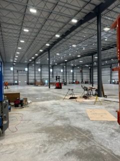 A wide, open warehouse interior with concrete floors, tall support columns, overhead lighting, and scattered equipment.