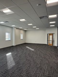 A vacant, brightly lit office space with light walls, patterned gray carpeting, recessed ceiling lights, and a glass door.
