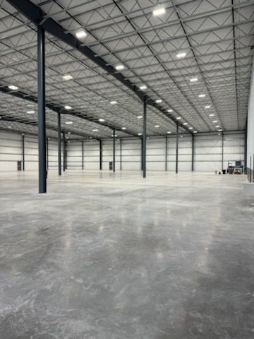 A large, empty industrial warehouse with high ceilings, concrete floors, steel support columns, and overhead LED lights.