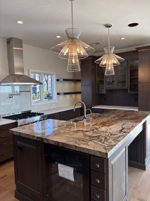 A kitchen island with a stone countertop, dark wooden cabinetry, and two tiered, light-colored pendant lights above.