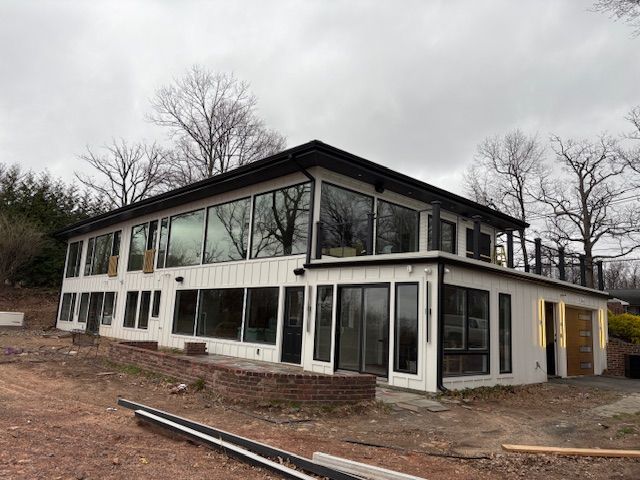 Modern two-story white house with expansive windows and a dark roof, set on a hillside surrounded by bare trees.