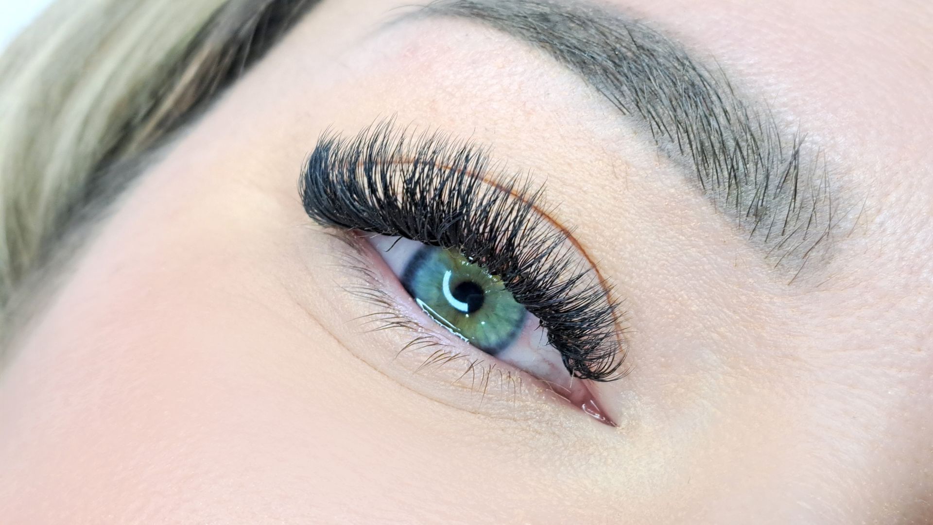 Close-up of a person's eye with long, dark lashes, a light brow, and bright green iris.