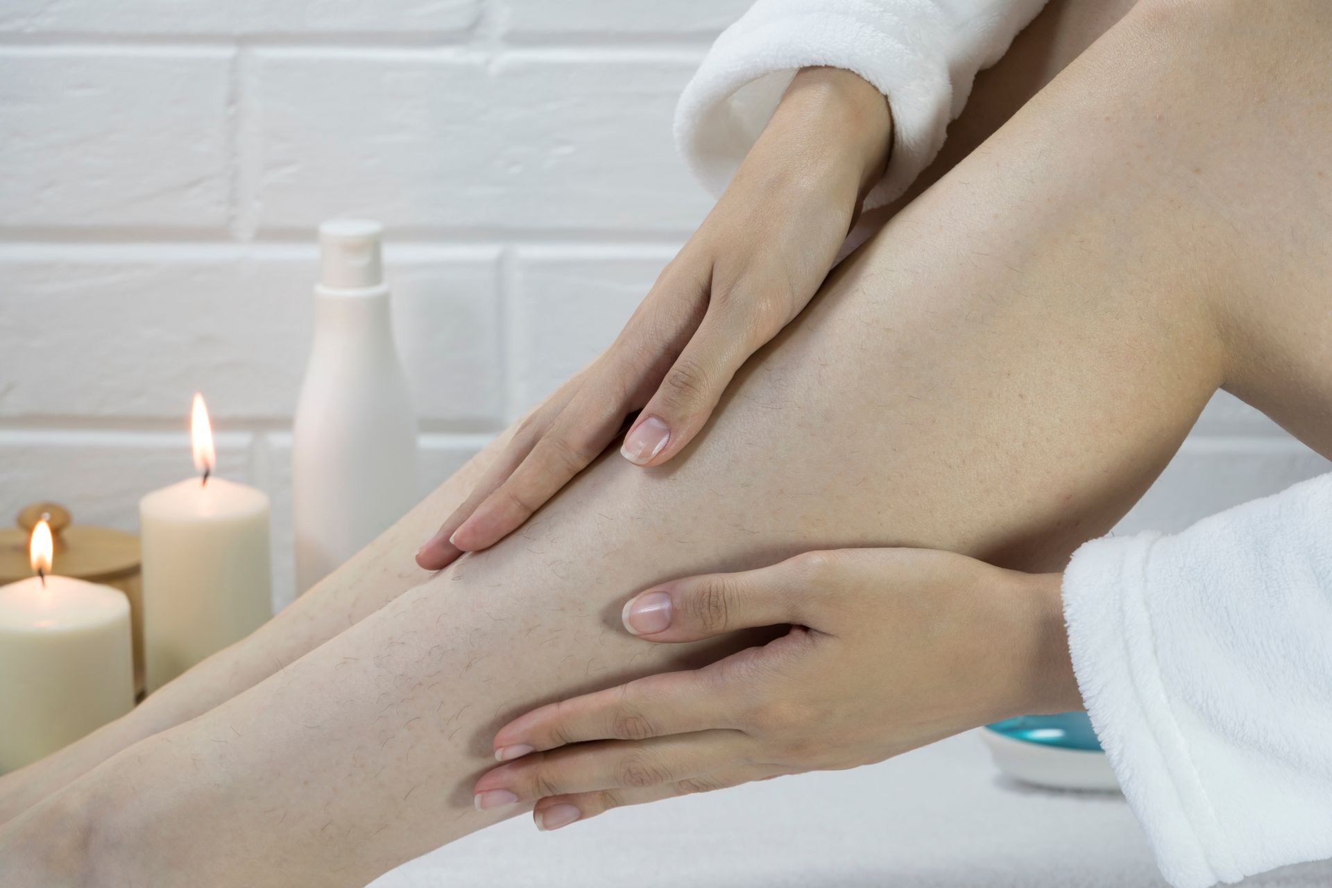 Woman's hands applying lotion to her leg, with candles and toiletries in a spa-like setting.