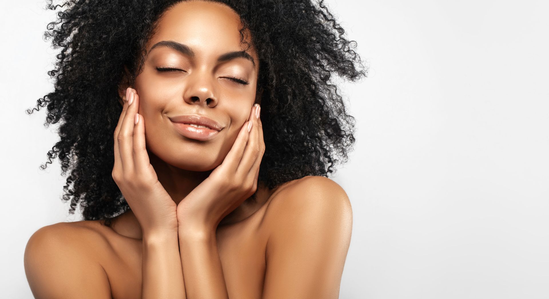 Woman with curly black hair, eyes closed, gently touching her face, against a white backdrop.