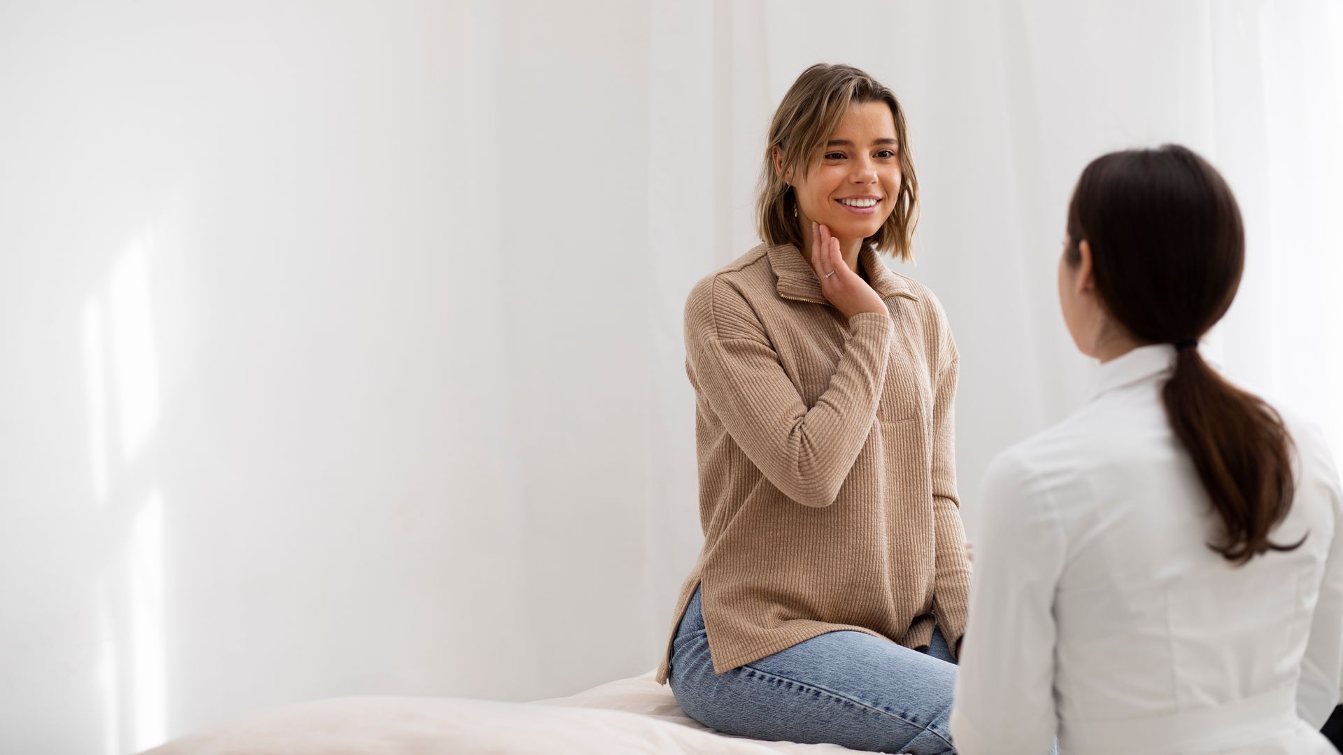 Woman in a tan sweater smiles while being examined by a medical professional in a white room.