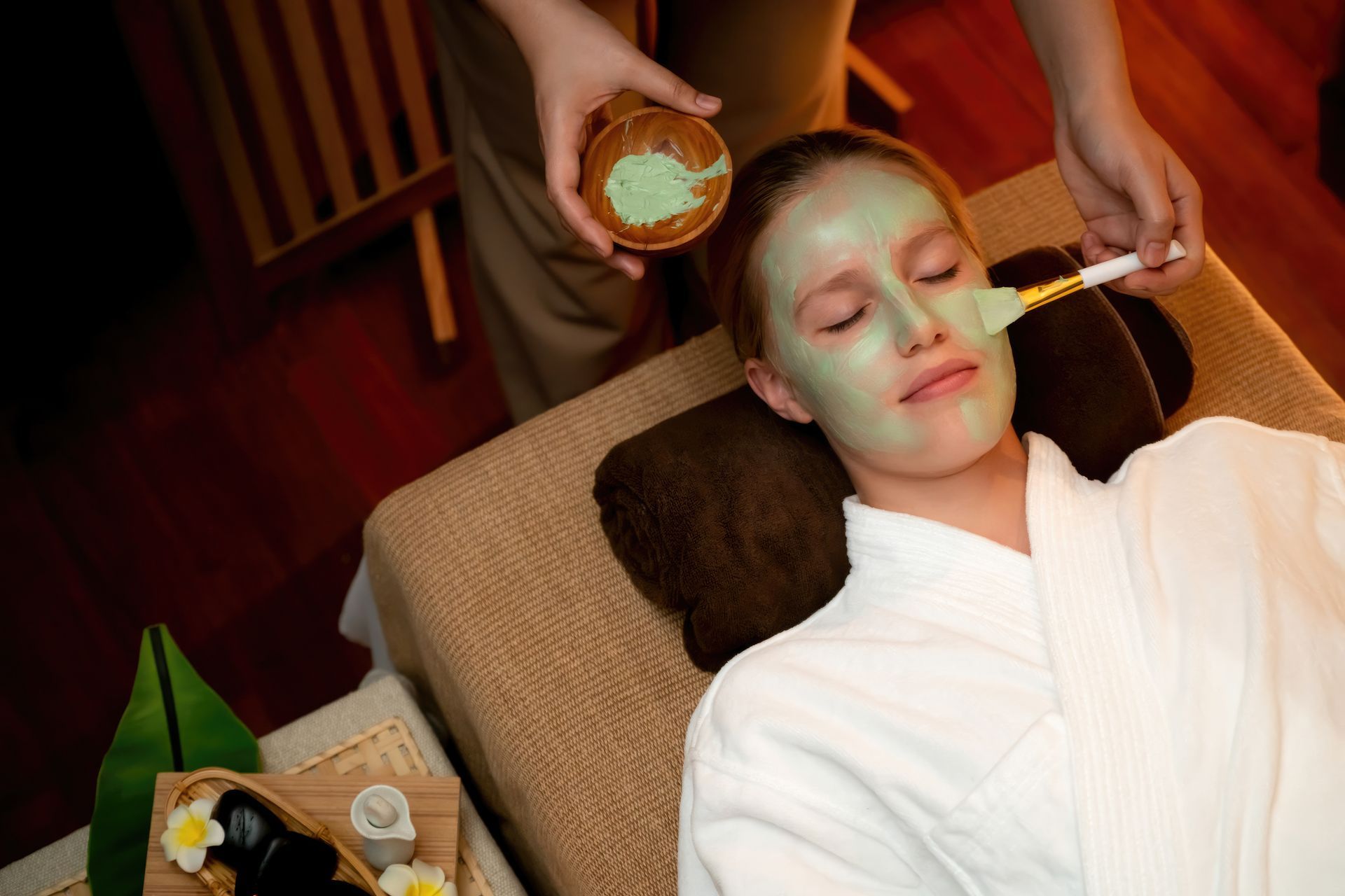 Woman receiving a facial mask treatment at a spa. A therapist applies a green mask to her face while she reclines in a bathrobe.