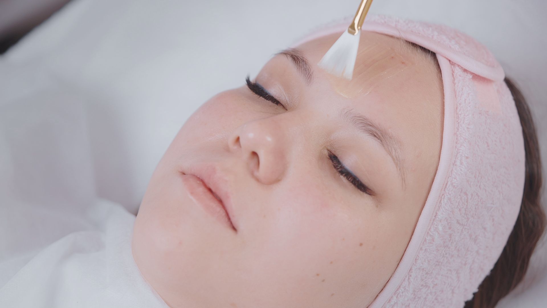 Woman receiving a facial treatment with a brush on her forehead, lying with eyes closed and a towel headband