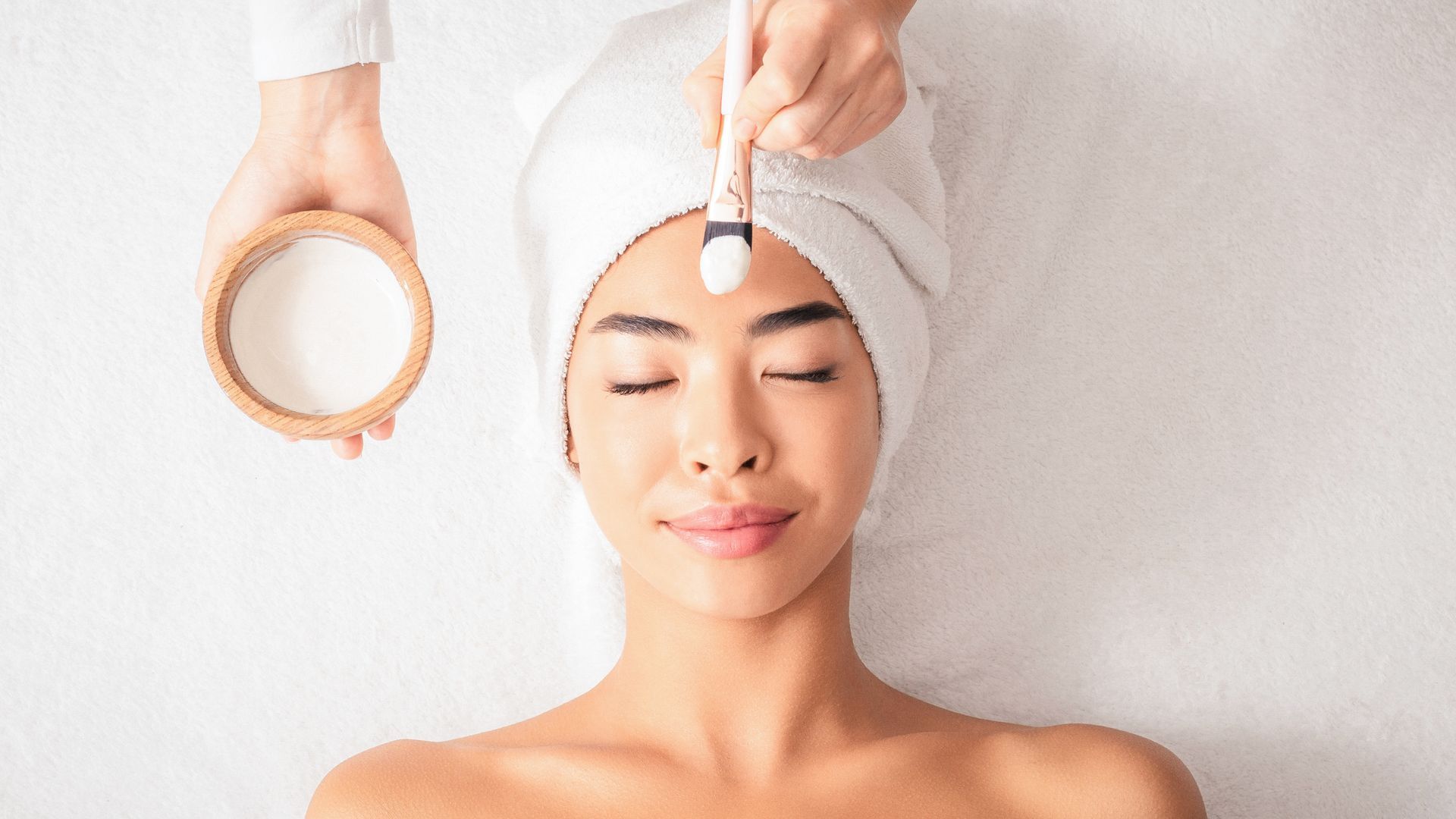Person receiving a facial treatment with a brush while lying on a bed, with a towel wrapped around the head