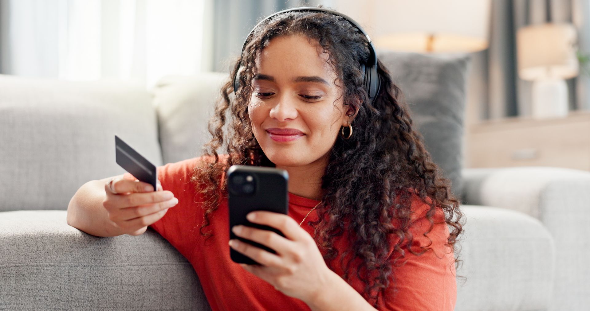 Person smiling on a couch, holding a phone and credit card while shopping online