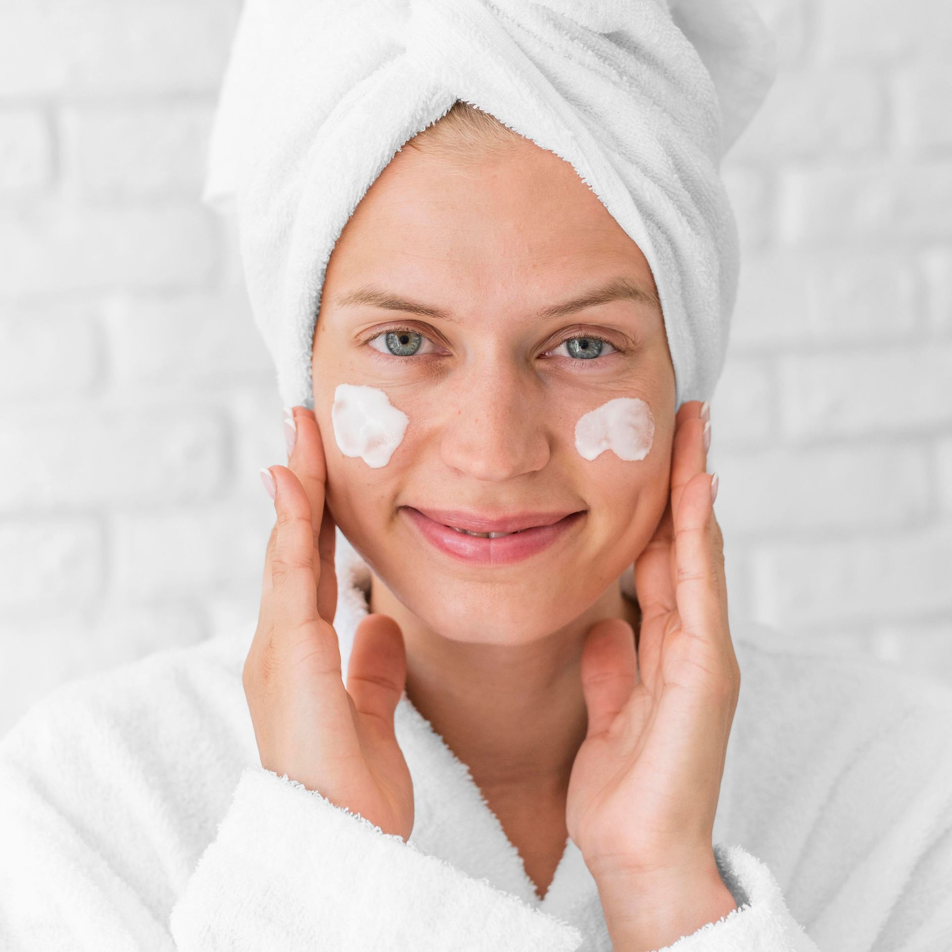 A woman with a towel wrapped around her head is applying lotion to her face.