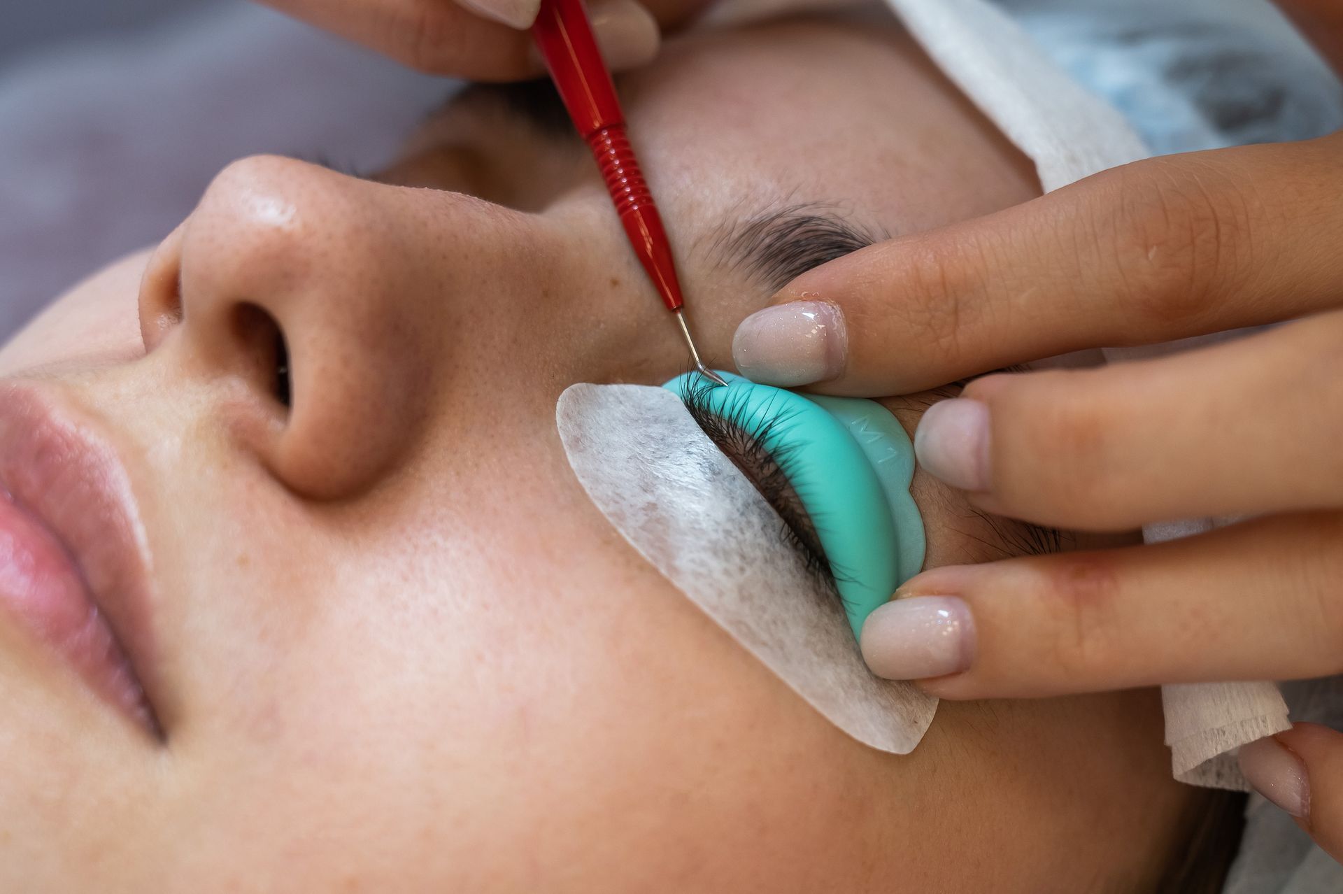 Person's eye during lash lift procedure; technician applying solution with a tool. Eye area with blue molds and white eye pads.
