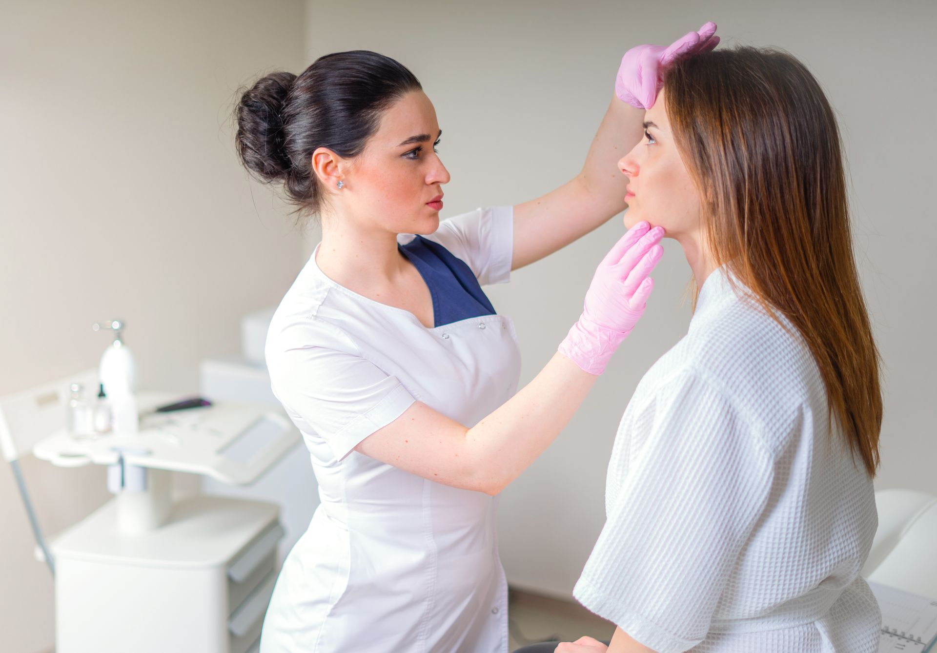 Cosmetologist examining a patient's face in a medical office, wearing gloves and white coat.