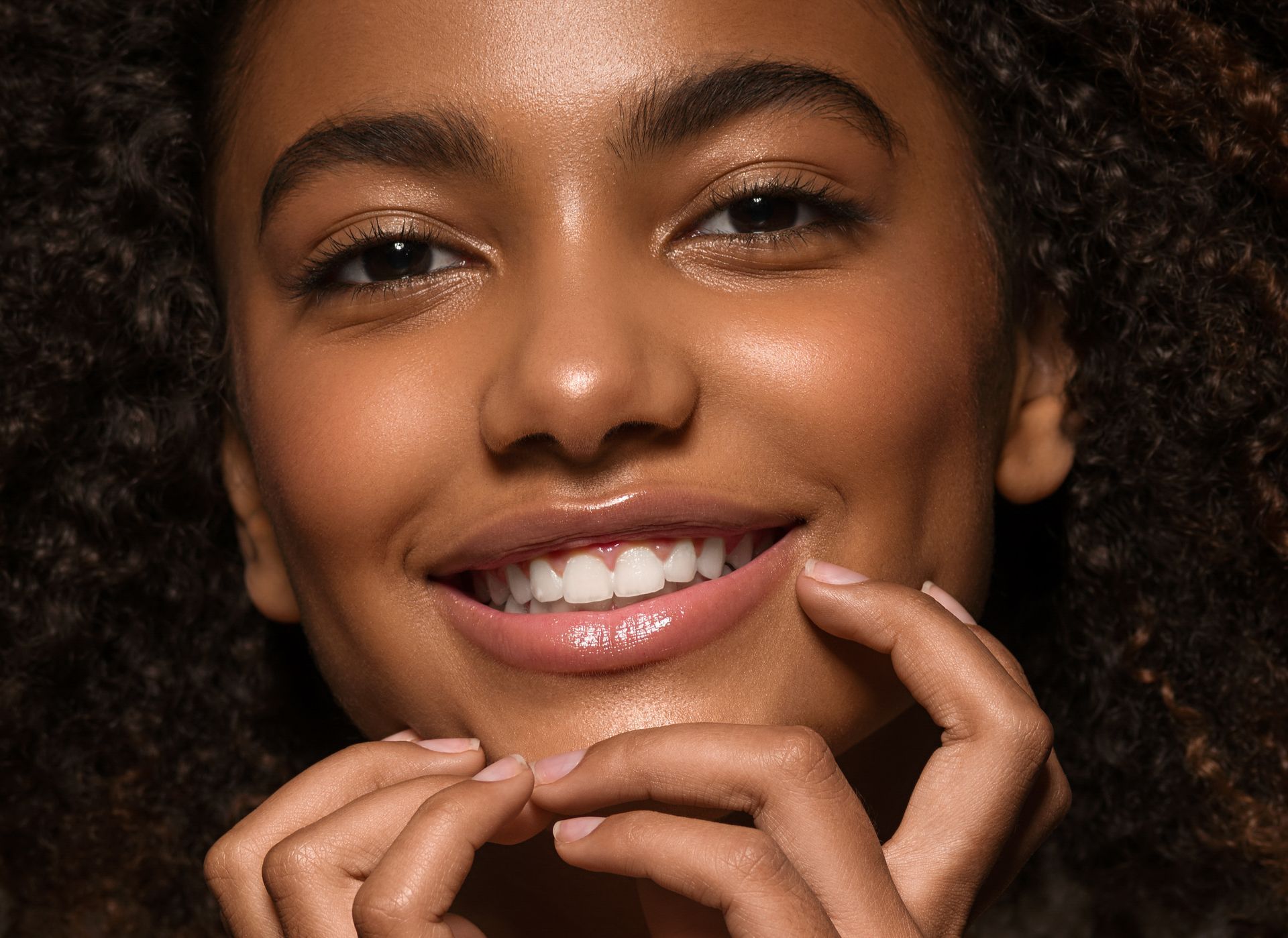 Close-up of a smiling dark-skinned woman with curly hair, touching her face with her fingers. She has bright eyes and white teeth.