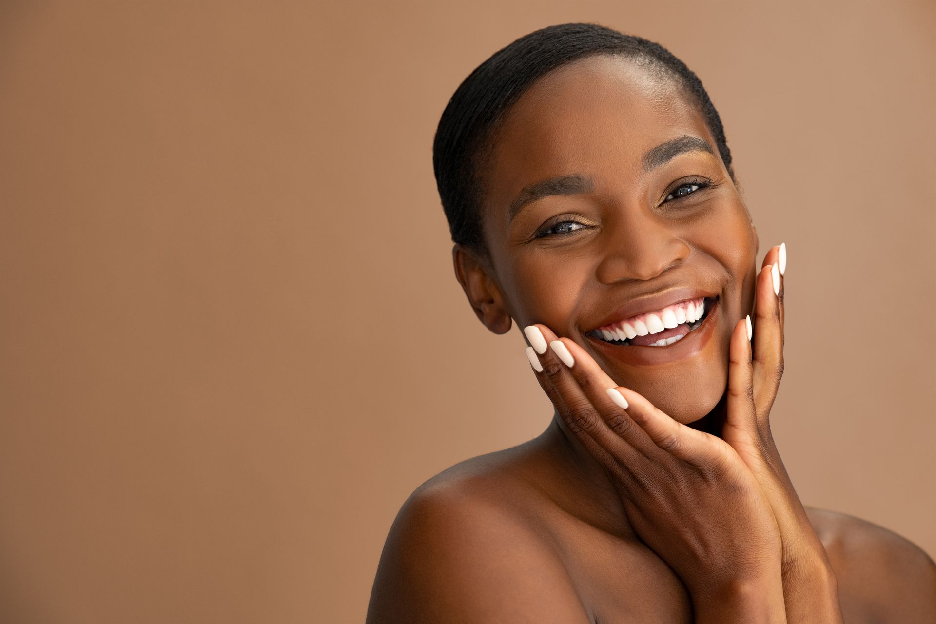 Woman with short hair, smiling broadly, holding hands up to cheeks, against a beige background.