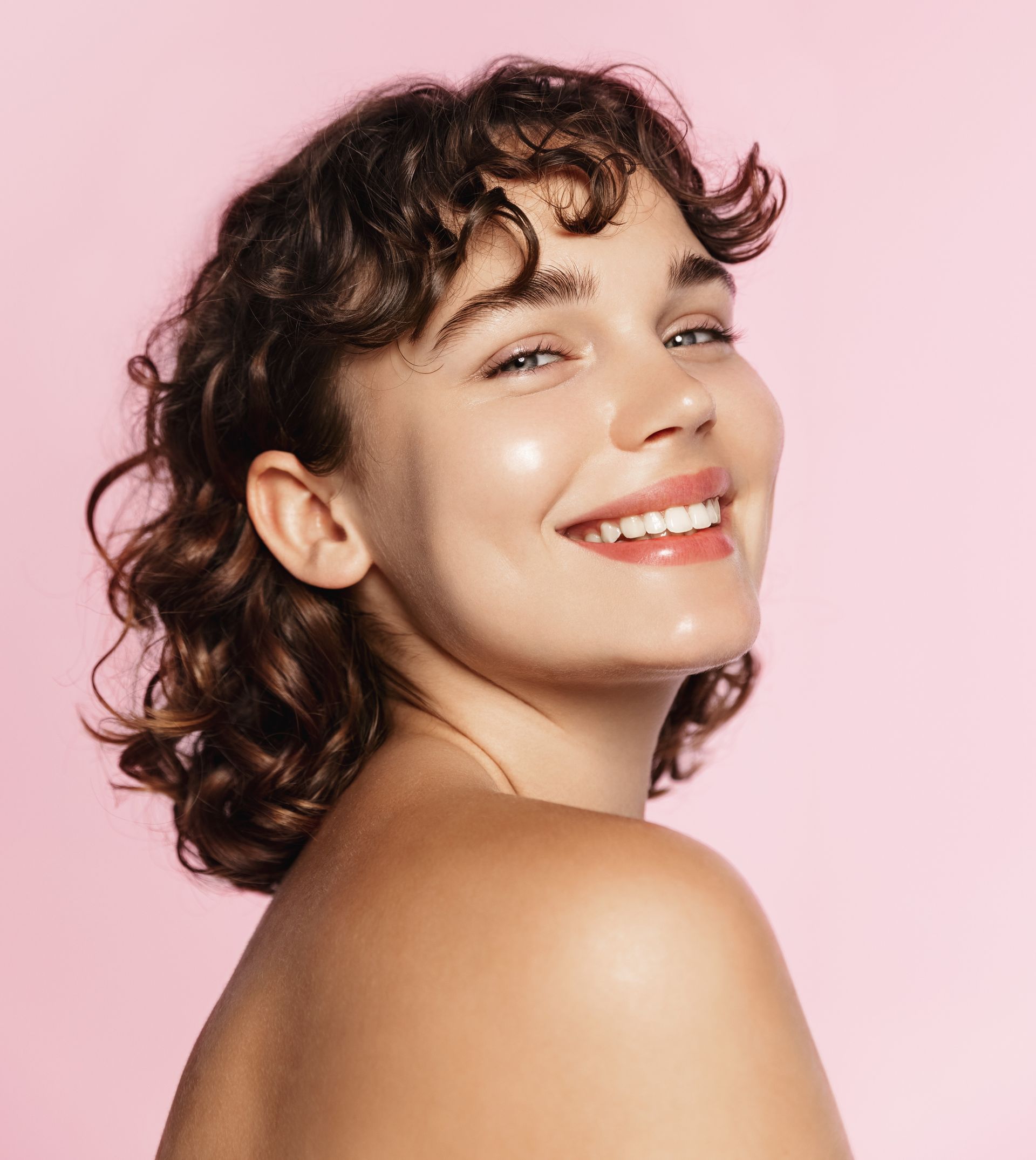 A woman with curly hair is smiling on a pink background.