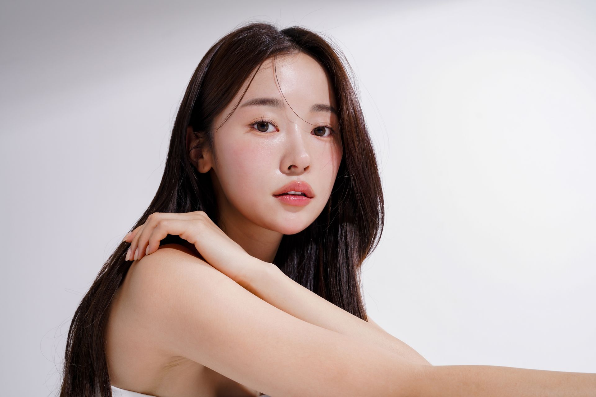 Woman with long dark hair, lightly smiling, resting arm on shoulder against white backdrop.