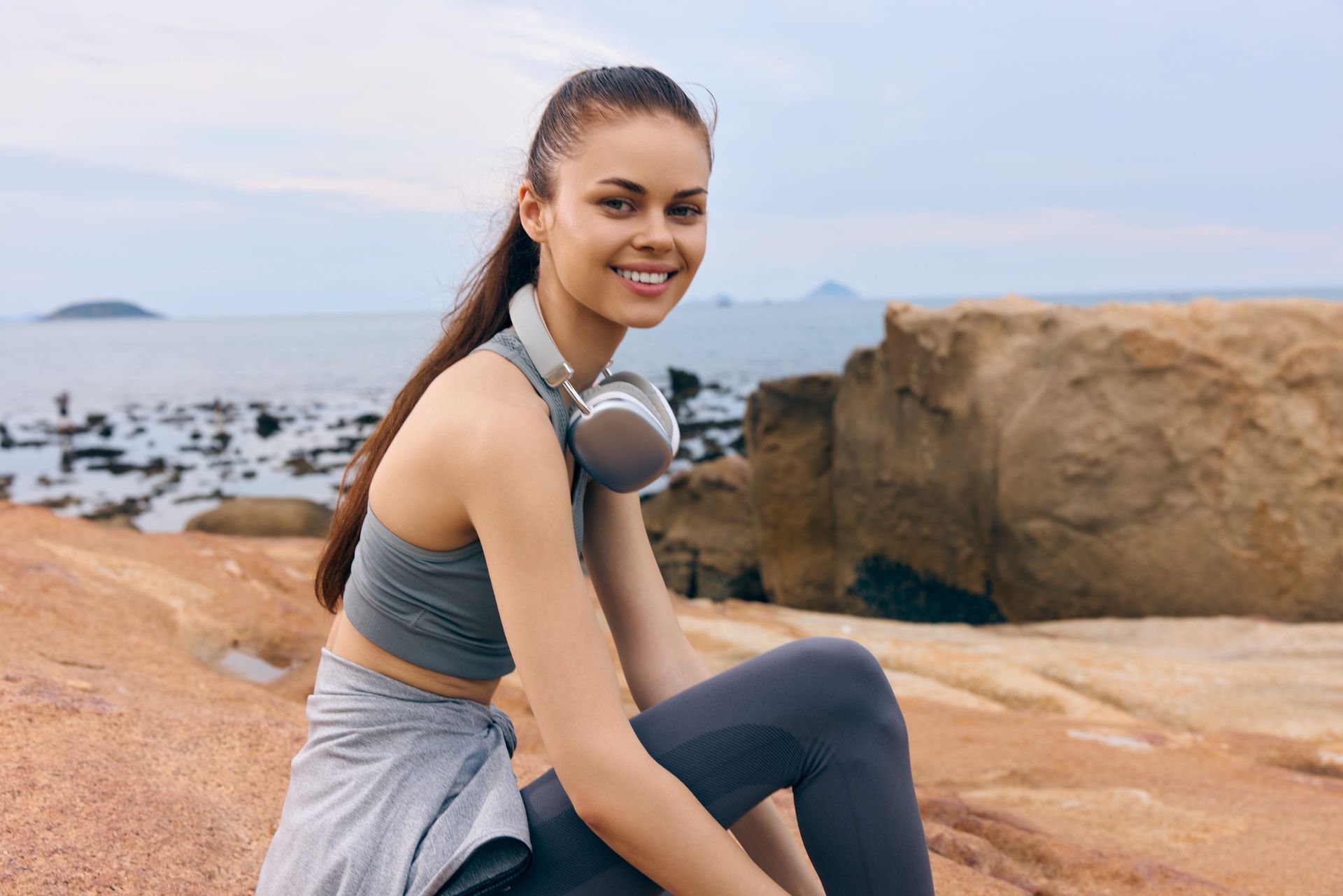 Woman in workout clothes smiles, sitting on rocks near the ocean, headphones around her neck.