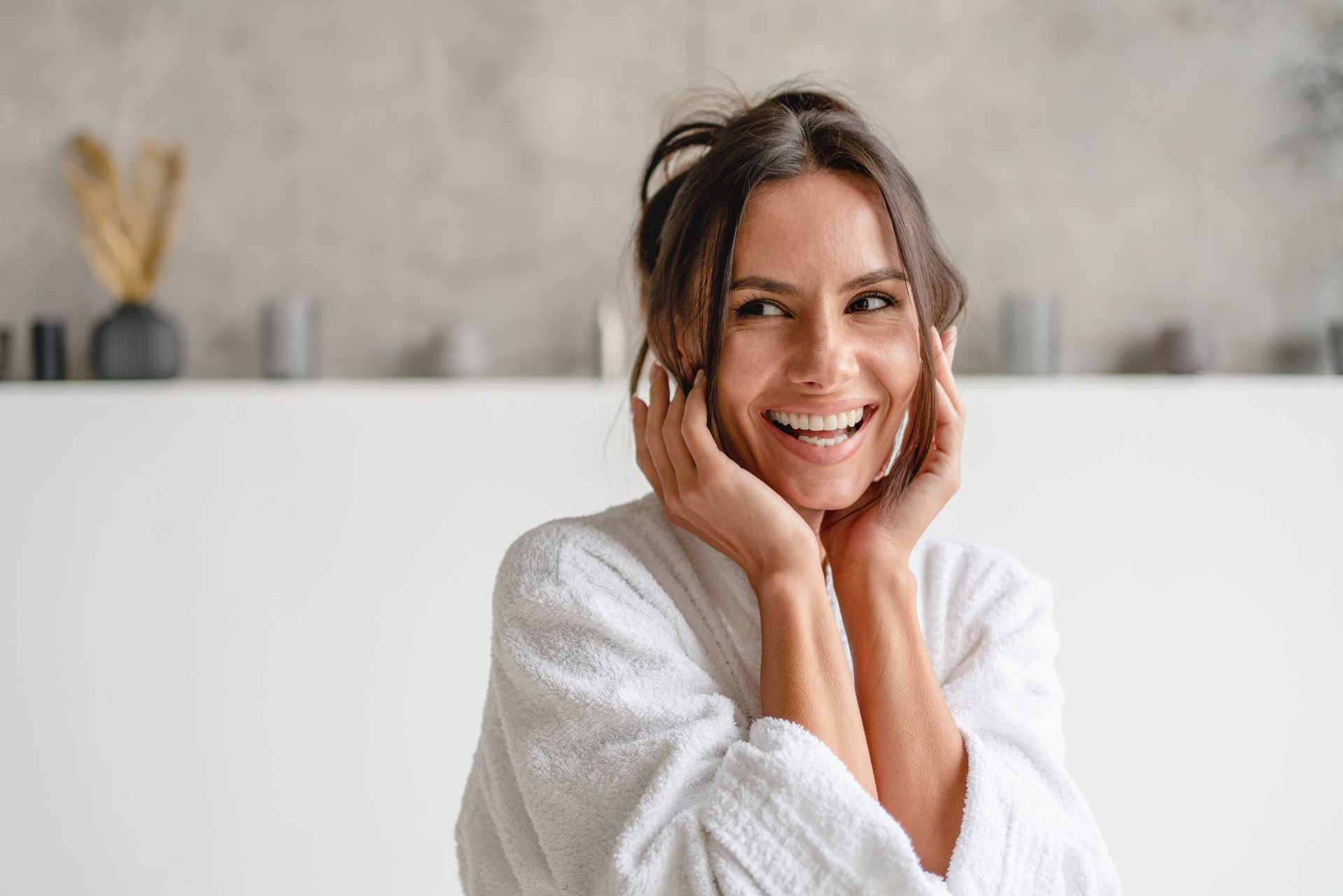 Woman in white bathrobe smiles, touching cheeks. Light, modern bathroom setting.