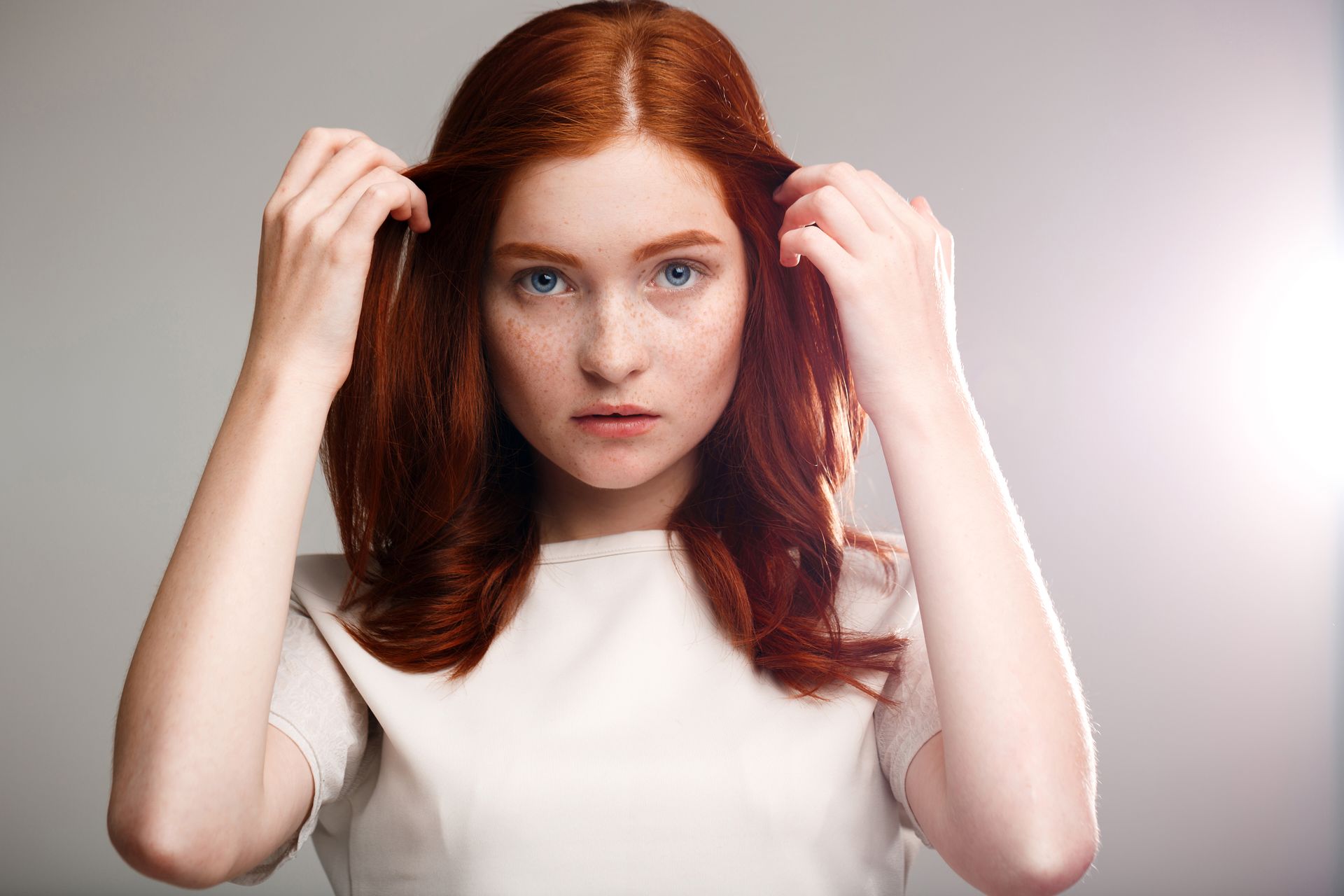 A woman with red hair and blue eyes is holding her hair.
