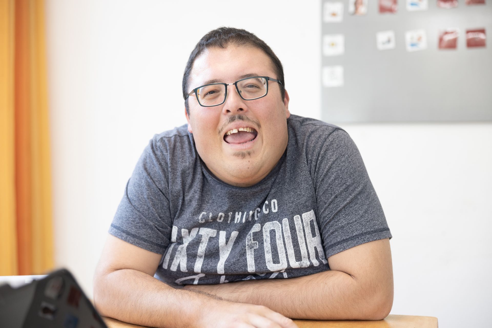 Ein Mann mit Brille und einem T-Shirt mit der Aufschrift „Sixty Four“ sitzt an einem Tisch.