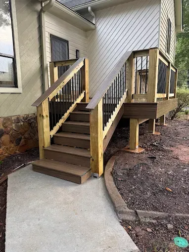 Wooden deck with stairs leading to a house. Brown and black railings with a concrete path.
