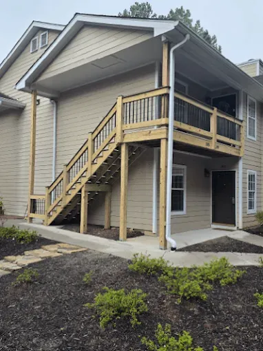 Exterior view of a two-story building with wooden stairs leading to a deck. Light tan siding, dark brown door, and black railings.