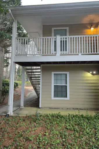 Two-story apartment building with exterior staircase and balcony. Tan siding, white railing, and windows.